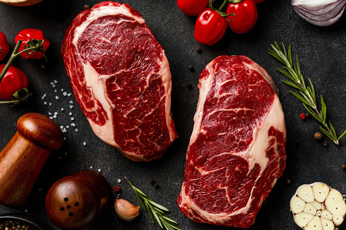 Two raw ribeye steaks on a dark surface with rosemary, garlic, and peppercorns.
