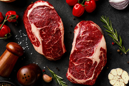 Two raw ribeye steaks on a dark surface with rosemary, garlic, and peppercorns.