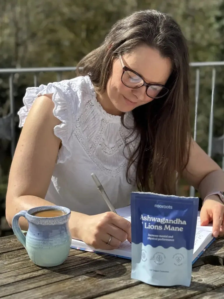 A woman writing at an outdoor table with her nooroots pouch and a mug